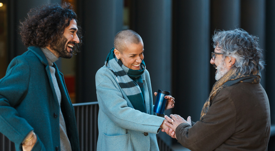 Three people in winter coats smile and shake hands while chatting outdoors.