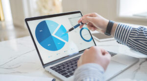 A person points at blue pie charts on a laptop screen while reviewing data at a desk.