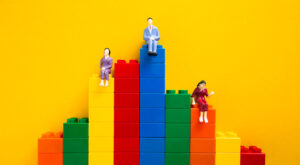 Miniature figurines sit on colorful stacked building blocks arranged in a bar graph pattern against a yellow background.